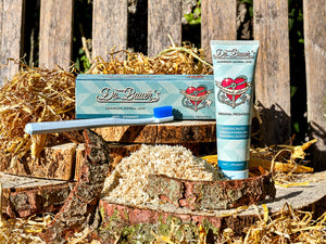 Dr. Bauer’s Zahnpasta ‚Mineral Love – Original Freshness‘ mit Verpackung und blauer Zahnbürste, arrangiert auf Holzscheiben, Stroh und Spänen vor einer rustikalen Holzbank – natürliche Inszenierung für remineralisierende Zahnpflege mit Meeresmineralien.