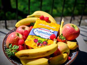 Fruchtige Szene mit Dr. Bauer’s Xylinetten-Packung in einer Schale voller Bananen, Äpfel, Erdbeeren und Himbeeren – natürliche Süße trifft auf Zahnpflege.
