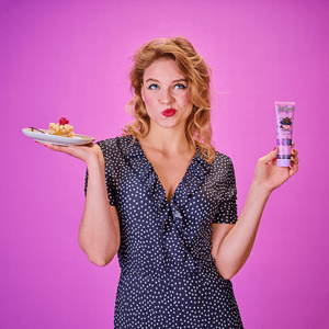Frau im gepunkteten Kleid vor pinkem Hintergrund hält einen Kuchenteller mit Himbeere in der einen und eine Dr. Bauer’s Zahnpasta ‚Fruits of Love Wild Love‘ in der anderen Hand – spielerischer Vergleich von Genuss und Zahnpflege.