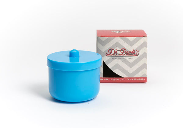 Eine Dr. Bauer's Reinigungsbad-Dose hellblau, ein blauer Plastikbehälter mit Deckel, steht neben einer rot-weißen Dr. Bauer's Box – perfekt zum Reinigen von Prothesen oder Zahnspangen.
