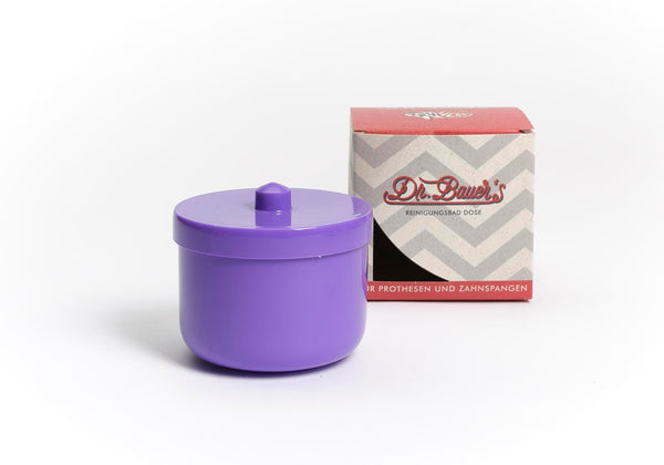 Die Dr. Bauer’s Reinigungsbad-Dose flieder/lila, ein violetter Behälter mit Deckel zur Zahnspangen- oder Prothesenreinigung, ist neben der Schachtel mit dem Dr. Bauer’s-Logo auf weißem Hintergrund abgebildet.