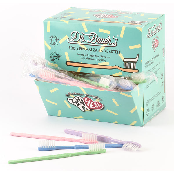 Vorne steht eine Schachtel Dr. Bauer's Einmalzahnbürsten -STROH- 100er MIX in violett, grün, blau und rot mit biologisch abbaubaren Einwegzahnbürsten mit integrierter Zahnpasta.