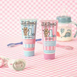 Zwei Tuben Babyzahnpasta und eine Babyflasche auf einem rosa karierten Untergrund mit Schnuller und Beißring.