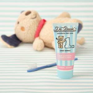 Dr. Bauer’s Baby-Zahnpasta ‚First Love – Sweet Berries‘ in pastellblauer Tube mit Teddybär-Motiv, daneben eine kleine Zahnbürste und im Hintergrund ein liegendes Kuscheltier – sanfte Szene für die erste Zahnpflege ohne Fluorid.