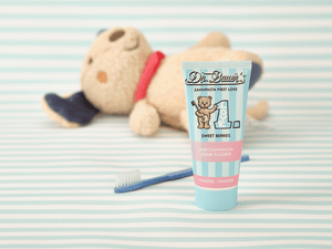 Dr. Bauer’s Baby-Zahnpasta ‚First Love – Sweet Berries‘ in pastellblauer Tube mit Teddybär-Motiv, daneben eine kleine Zahnbürste und im Hintergrund ein liegendes Kuscheltier – sanfte Szene für die erste Zahnpflege ohne Fluorid.