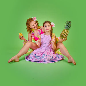 Fröhliche Frau im Hawaii-Outfit sitzt barfuß hinter einem Mädchen im rosa Kleid, das eine Zahnbürste und eine Tube Dr. Bauer’s Zahnpasta ‚Fruits of Love – Fruit Splash‘ hält; im Hintergrund Ananas und Orange – tropisch-bunter Look für kindgerechte Zahnpflege.