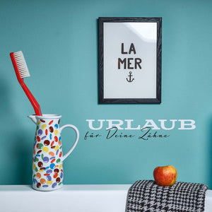 Große Zahnbürste in buntem Krug, Apfel auf Handtuch, gerahmtes ‚La Mer‘-Bild an türkisfarbener Wand, daneben der Schriftzug ‚Urlaub für Deine Zähne‘ – Szene im Badezimmer-Stil.“