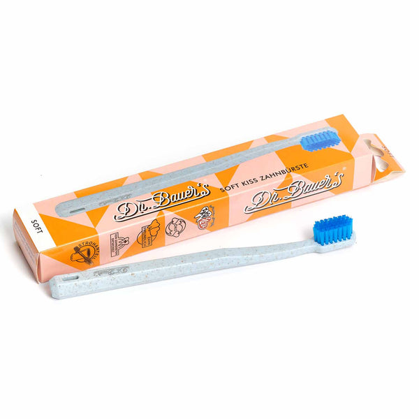Eine hellblaue Dr. Bauer‘s Soft Kiss Zahnbürste soft mit blauen biobasierten Borsten steht neben ihrer orange-weißen Dr. Bauer‘s-Box.