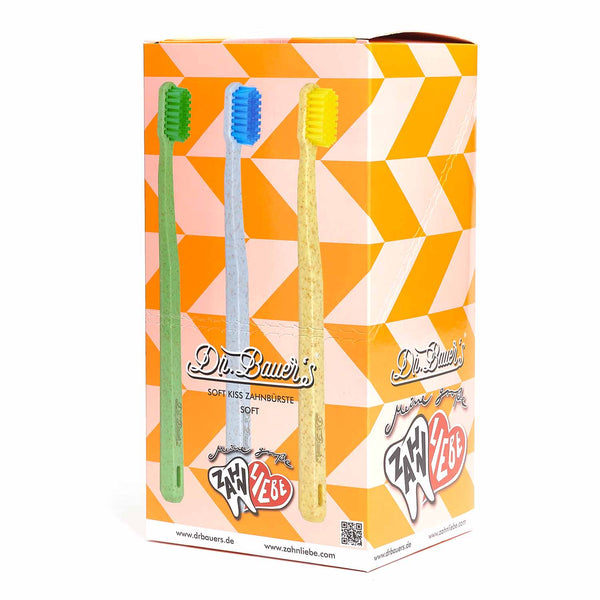 Box mit drei bunten Dr. Bauer's Soft Kiss Zahnbürsten soft (grün, blau, gelb) auf orangefarbenem geometrischem Hintergrund – jede Zahnbürste ist aus umweltfreundlichen Materialien hergestellt.