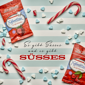 Zwei Packungen Dr. Bauers Xylinetten-Bonbons sind auf einer blau-weiß gestreiften Fläche ausgestellt, umgeben von Zuckerstangen und kleinen herzförmigen Bonbons. Der deutsche Text befindet sich in der Mitte.