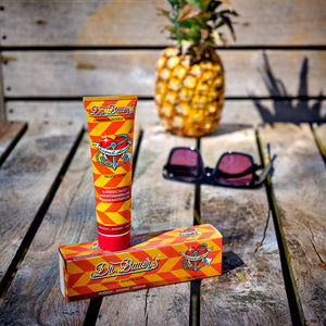 Dr. Bauer’s Zahnpasta ‚Mineral Love – Tropic Love‘ mit Verpackung auf einer Holzbank inszeniert; im Hintergrund eine Ananas und eine Sonnenbrille – sommerliches Setting für remineralisierende Zahnpflege mit tropischem Geschmack.