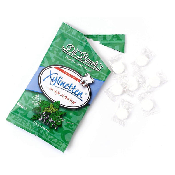 Abgebildet ist ein grüner Beutel Dr. Bauer's Xylinetten Pfefferminz 60g, ideal für die Zahnpflege und Kariesprophylaxe, daneben mehrere einzeln verpackte weiße Mints.