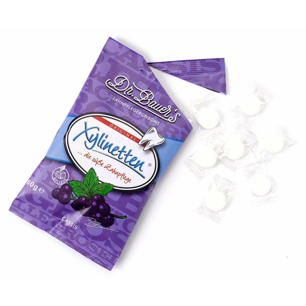 Dr. Bauer's Xylinetten Cassis 60g-Beutel mit daneben verstreuten weißen runden Zahnpflege-Bonbons, aromatisiert mit schwarzer Johannisbeere.