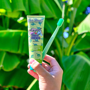 Eine Hand hält eine grüne Zahnpastatube und eine grüne Zahnbürste vor großen grünen Blättern im Freien.