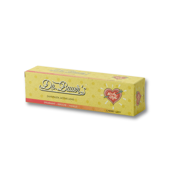 Eine gelbe Dr. Bauer's Schachtel zeigt "Zahnpasta Active Love Cherry Lady 90ml" mit einem herzförmigen "Apple Love"-Logo und wirbt für ayurvedische Zahnpflege.