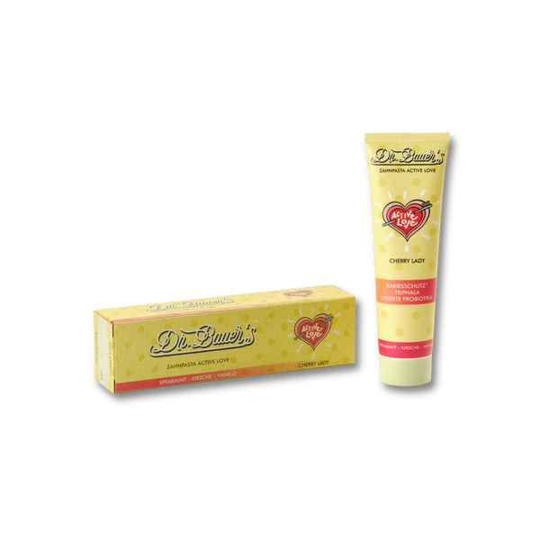 Eine gelbe Tube von Dr. Bauer's Zahnpasta Active Love Cherry Lady 90ml mit Probiotikum steht neben der dazugehörigen Schachtel, beide mit rotem Herzlogo.