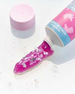 Aus einer Tube wird leuchtend pinkes Gel mit silbernem Glitter auf eine weiße Fläche gepresst; daneben liegt eine pinke Kappe.