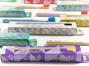 Dr. Bauer‘s Hero Love Whitening Zahnbürste und Boxen, mit farbenfrohen Designs und Supersoftborsten für eine sanfte Reinigung, angeordnet auf einer weißen Oberfläche.