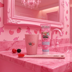 In einem rosafarbenen Badezimmer mit Einhorn- und Erdbeerdekor stehen eine rosafarbene Zahnbürste, ein Becher und Dr. Bauer's Lovely Friends Einhorn Kinderzahnpasta mit Glitzer auf dem Tresen und verleihen dem Raum verspielte Farbe und Charme.