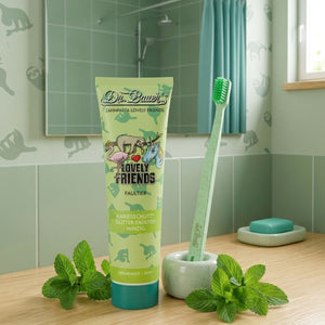 Dr. Bauer's Zahnpasta Lovely Friends Faultier 90ml Tube, verziert mit niedlichen Tierillustrationen, steht zusammen mit einer grünen Zahnbürste auf dem Waschbecken, Minzblätter in der Nähe verstreut. Marke: Dr. Bauer's.