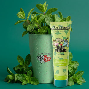 Eine Tube Dr. Bauer's Zahnpasta Lovely Friends Faultier 90ml steht neben einer mit Minze gefüllten Tasse auf grünem Hintergrund; die Produktbeschreibung zeigt nur den CSS-Code, ohne eigentliche Produktinformation oder Text.