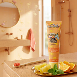 Auf dem Waschbecken steht eine Tube Dr. Bauer's Flamingo Zahnpasta Kinder - Lovely Friends (90 ml, ab 6 Jahren, mit Xylitol) im sauberen Helvetica Neue-Stil, daneben Zitrusfrüchte und eine Zahnbürste.