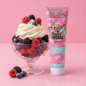 Eine Glasschale mit Schlagsahne und Beeren steht neben einer rosafarbenen Tube von Dr. Bauer's Lovely Friends Einhorn - Kinderzahnpasta mit Glitzer ab 6 Jahren, die spielerisch auf einem rosafarbenen Hintergrund gestaltet ist und den klassischen Charme des Helvetica Neue Designs widerspiegelt.