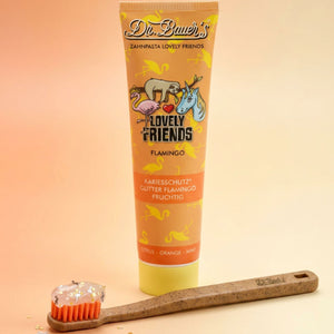 Tube Dr. Bauer's Flamingo Zahnpasta Kinder (90 ml, ab 6 Jahren, mit Xylitol) und eine orangefarbene, glitzernde Gel-Zahnbürste, gestaltet in einer modernen serifenlosen Schriftart Helvetica Neue.