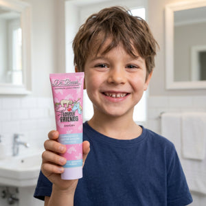 Ein lächelnder Junge im blauen Hemd hält in einem hellen Badezimmer Dr. Bauer's Lovely Friends Einhorn - Kinderzahnpasta mit Glitzer ab 6 Jahren, eine glitzernde Kinderzahnpasta von Dr. Bauer's, hoch. Es ist keine Produktbeschreibung enthalten - nur CSS-Code für Styling-Elemente.