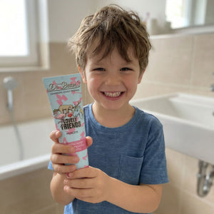 Ein lächelndes Kind in einem blauen Hemd hält die Zahnpasta Lovely Friends Prinzessin 90ml von Dr. Bauer in der Hand. Sie spiegelt die klaren, modernen Linien des von der Helvetica Neue inspirierten Designs in einem hellen Badezimmer wider.
