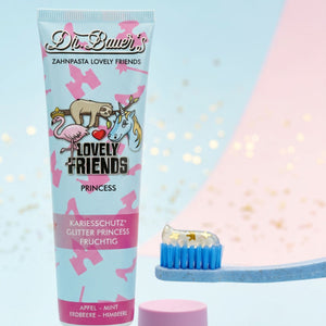 Eine Tube Dr. Bauer's Zahnpasta Lovely Friends Prinzessin 90ml und eine blaue Zahnbürste mit glitzernder Zahnpasta.