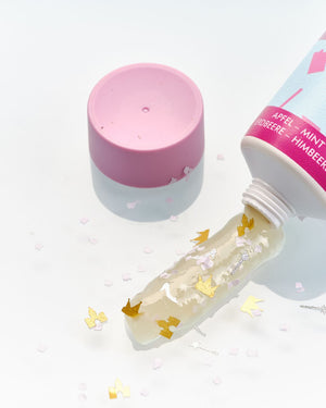 Aus einer pastellfarbenen Tube mit rosafarbener Kappe tropft ein mit goldenem und weißem Konfetti verziertes Gel auf eine weiße Fläche.