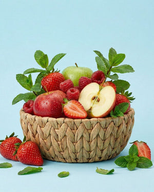 Ein geflochtener Korb, gefüllt mit Äpfeln, Erdbeeren, Himbeeren und Minzblättern auf einem hellblauen Hintergrund.