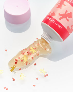 Klares Gel mit goldenem und rotem Konfetti in Bärenform, das aus einer rosafarbenen Tube mit der Aufschrift Erdbeere-Mint herausquillt.