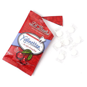 Eine rote Tüte mit Dr. Bauer‘s Xylinetten Kirsche 60g Bonbons, die für Zahngesundheit und Kirschgenuss werben, aus der mehrere weiß verpackte Xylinetten der Marke Dr. Bauer‘s herausquollen.