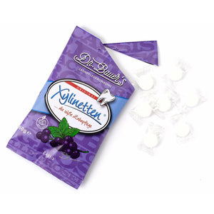 Dr. Bauer's Xylinetten Cassis 60g-Beutel mit daneben verstreuten weißen runden Zahnpflege-Bonbons, aromatisiert mit schwarzer Johannisbeere.